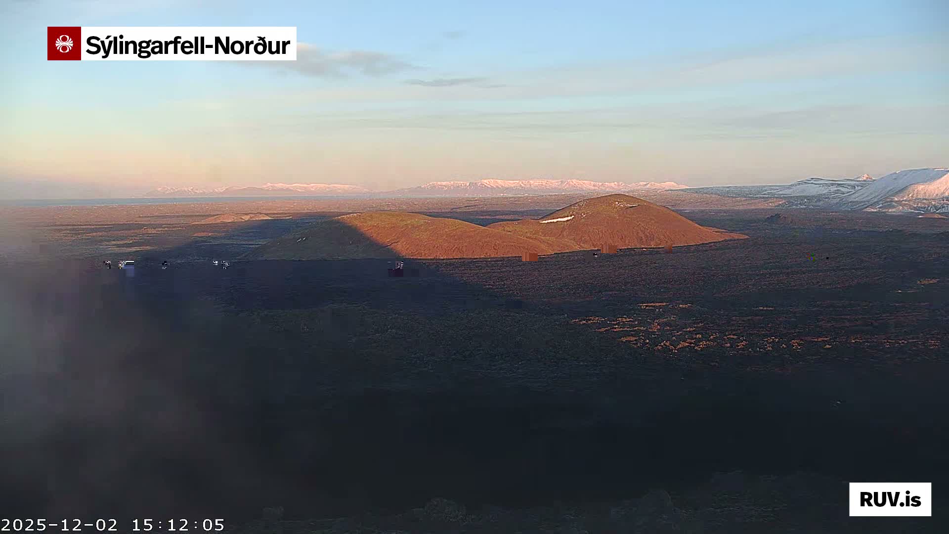 A vast, rugged volcanic landscape is seen under clear, sunny skies, featuring two prominent, sun-drenched conical hills, several small structures, and a backdrop of distant snow-capped mountains.