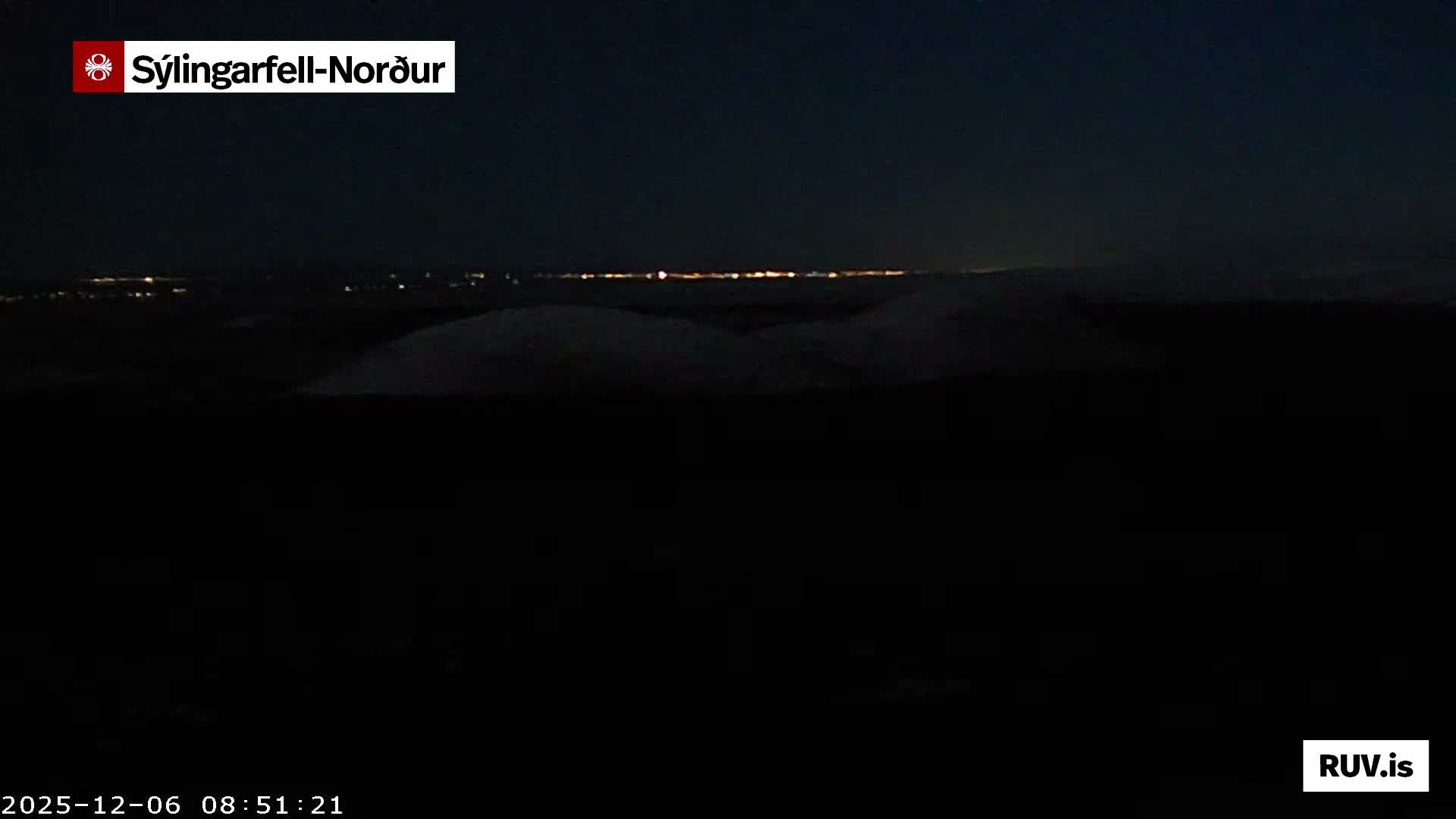 A dark nighttime landscape reveals undulating, possibly snow-covered terrain in the foreground with a distant band of city lights under a clear sky.