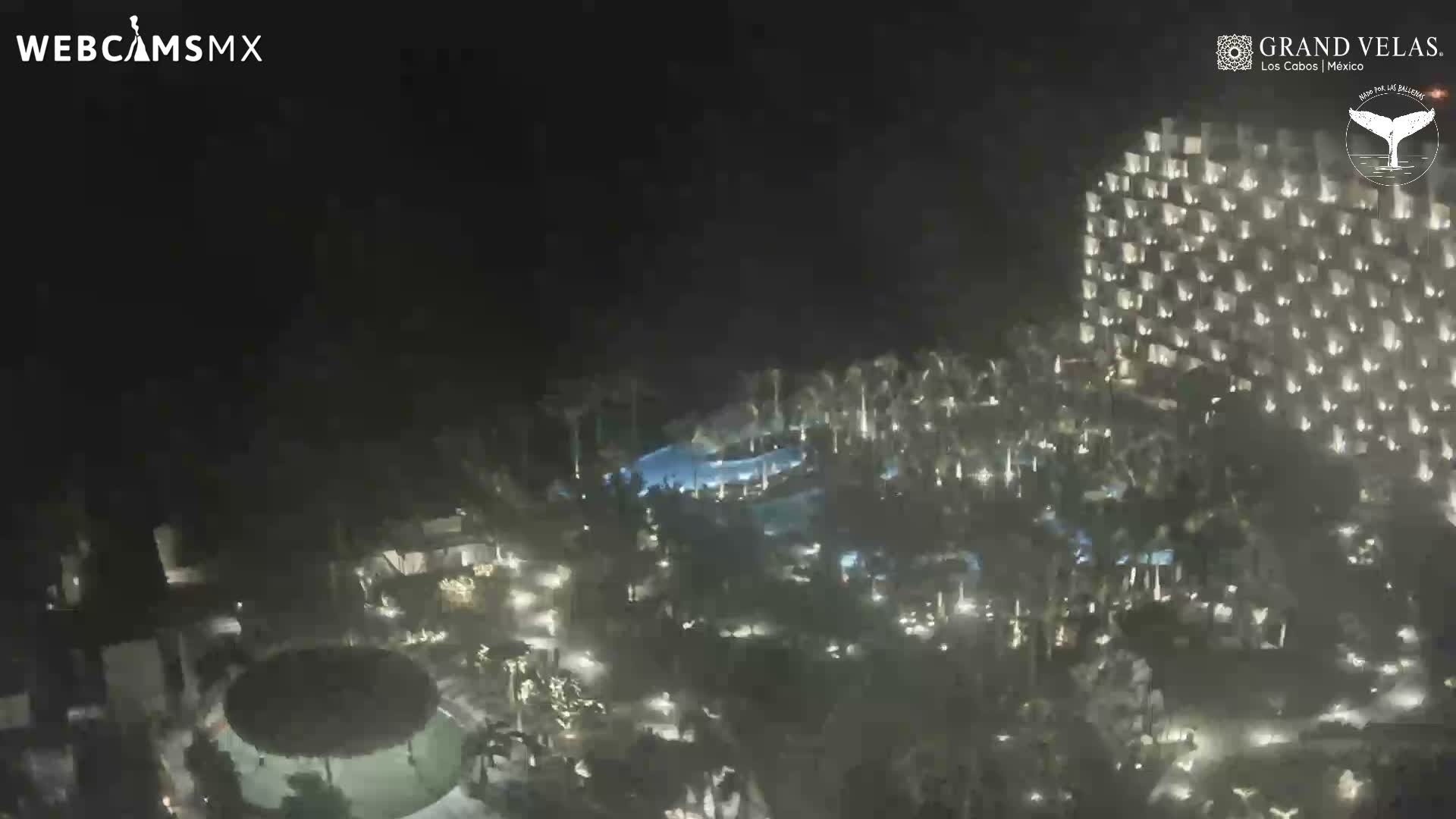 A sprawling resort complex is brightly illuminated at night, featuring a large multi-story hotel building, multiple glowing blue swimming pools, and numerous palm trees under a clear dark sky.