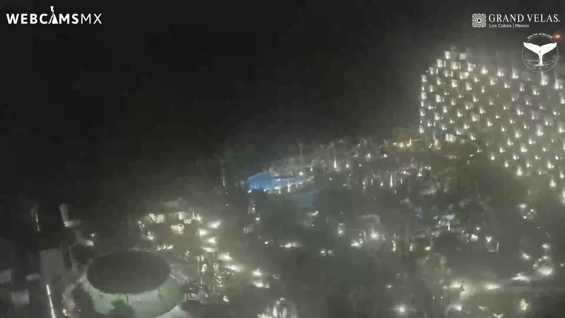 A sprawling resort complex is brightly illuminated at night, featuring a large multi-story hotel building, multiple glowing blue swimming pools, and numerous palm trees under a clear dark sky.