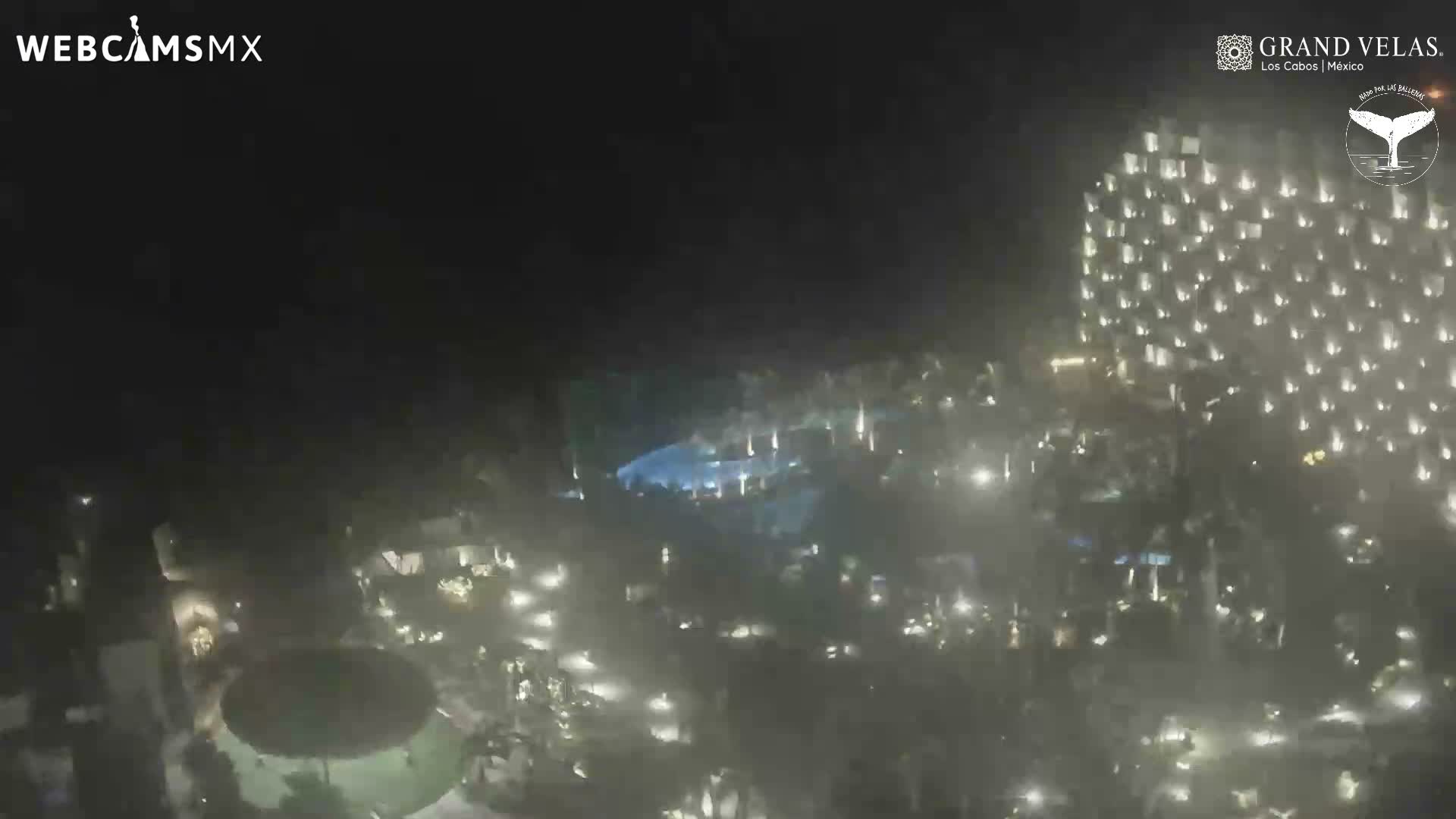 An aerial night view shows a brightly illuminated resort complex with a tall, light-dotted building, multiple glowing swimming pools, and extensive outdoor areas under a mildly hazy sky.