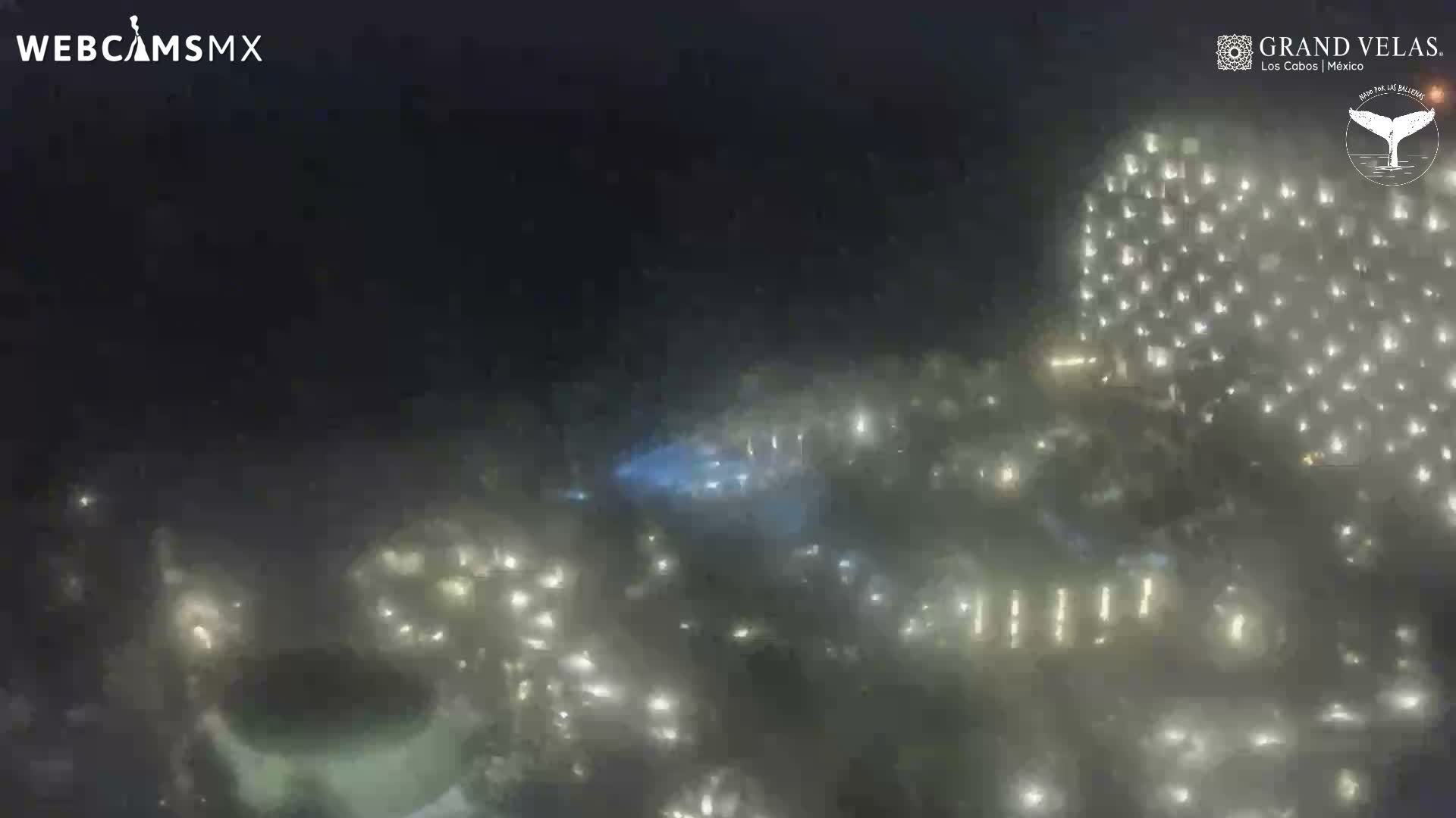 A nighttime outdoor scene captures a large building brightly illuminated by countless white lights and a central area with blue lights, all appearing diffused through a misty or hazy atmosphere.