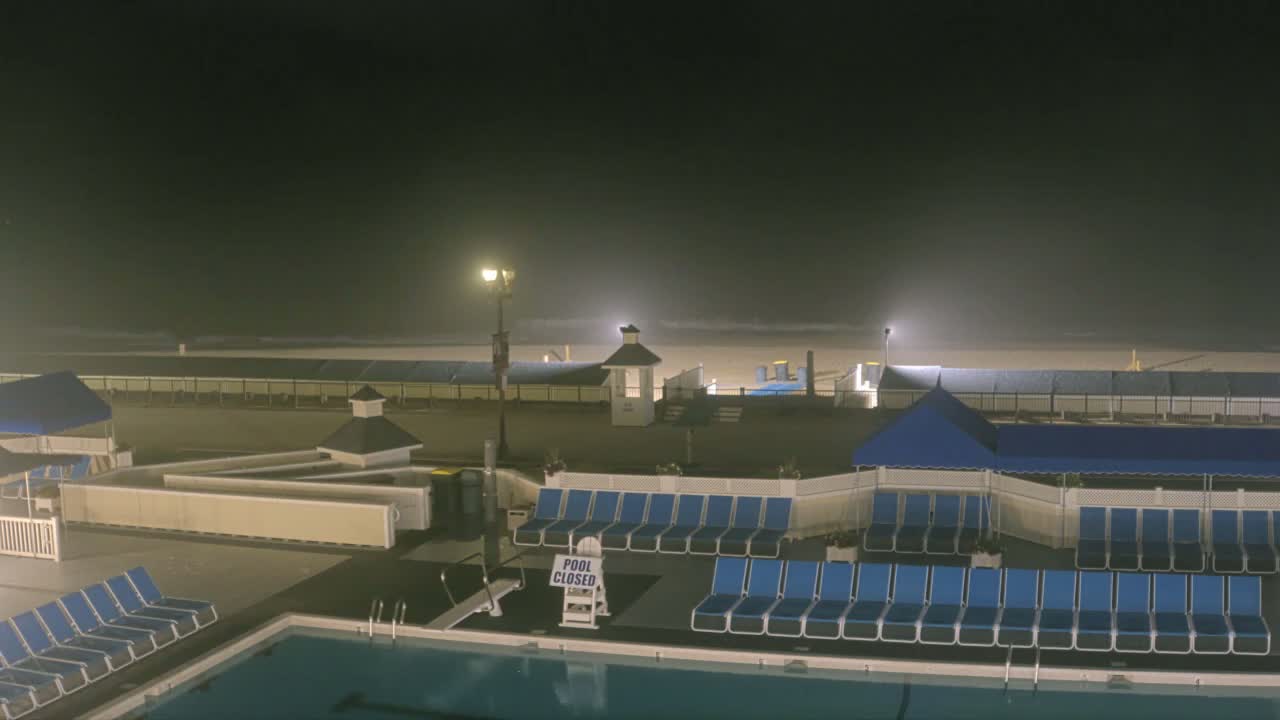 A nighttime view of a beachside pool area with rows of empty lounge chairs, a closed pool, and a foggy beach in the background.