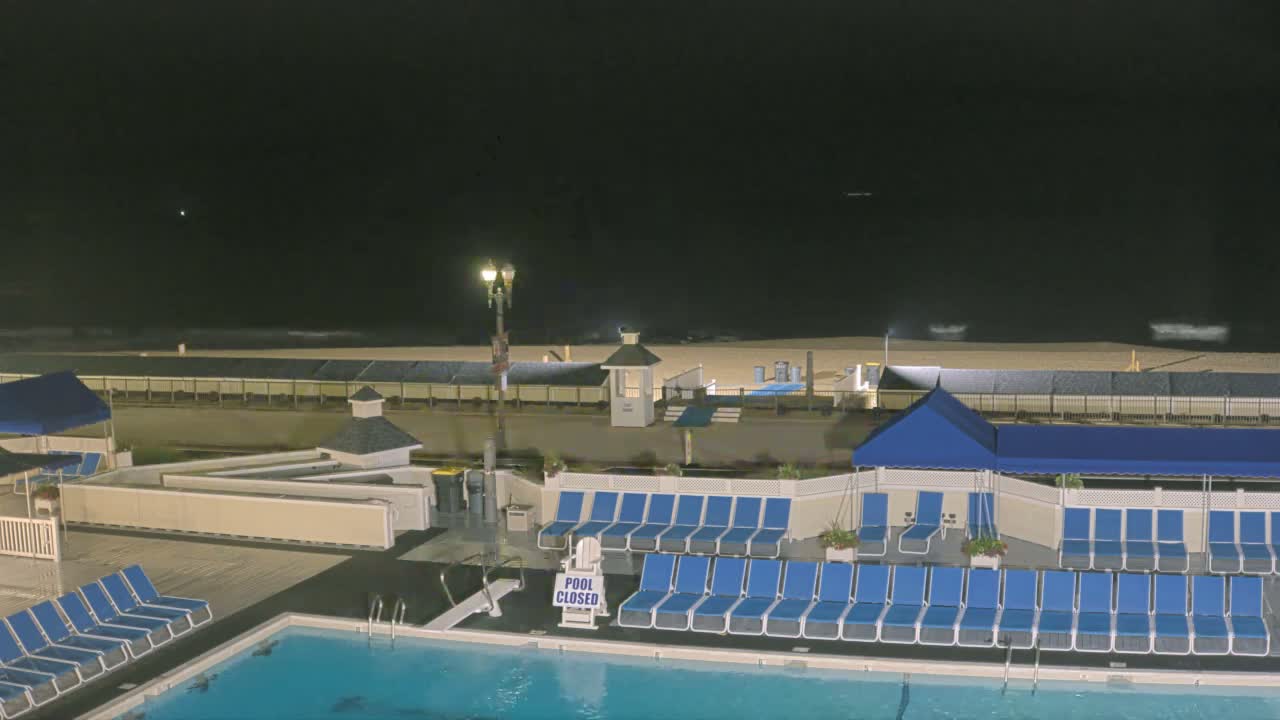 An outdoor nighttime scene shows a closed swimming pool surrounded by rows of blue lounge chairs, a beach beyond a fence, and a dark, somewhat overcast sky.