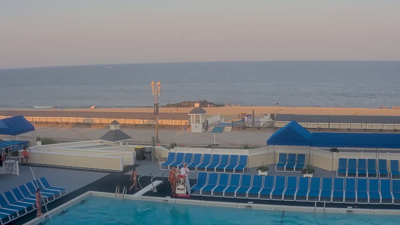 An outdoor nighttime scene shows a closed swimming pool surrounded by rows of blue lounge chairs, a beach beyond a fence, and a dark, somewhat overcast sky.