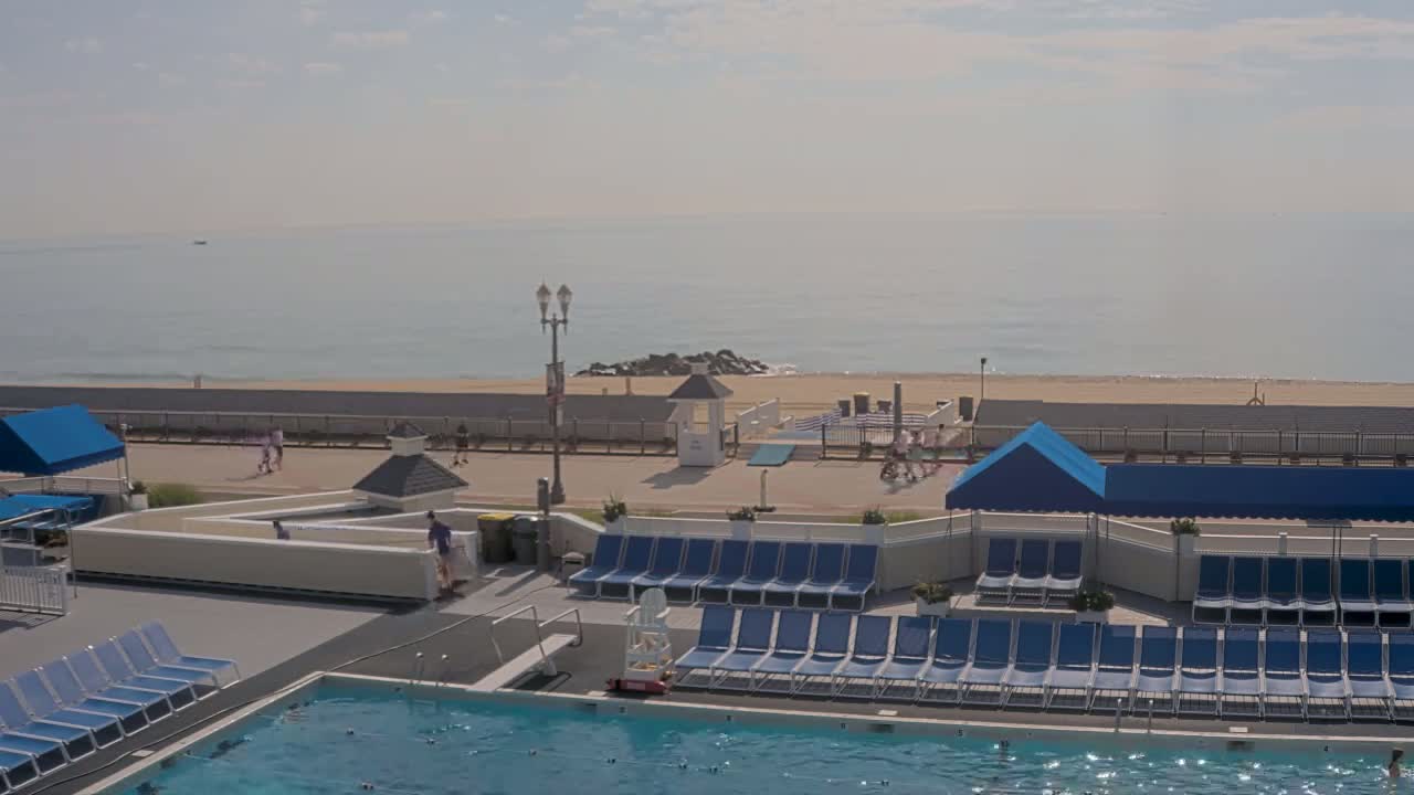 An outdoor nighttime scene shows a closed swimming pool surrounded by rows of blue lounge chairs, a beach beyond a fence, and a dark, somewhat overcast sky.
