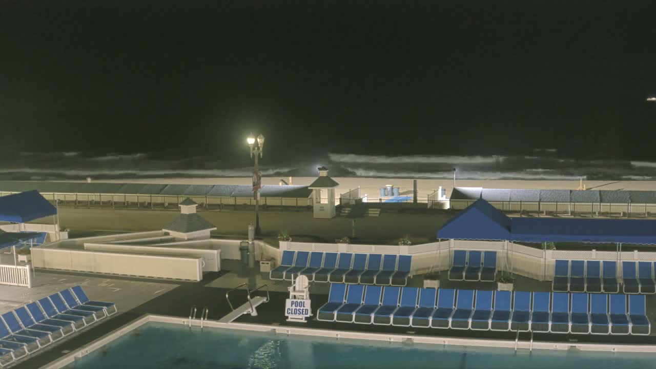 A nighttime view of a beach with moderately rough waves, showing a closed pool area with rows of blue lounge chairs and a lifeguard chair next to it.