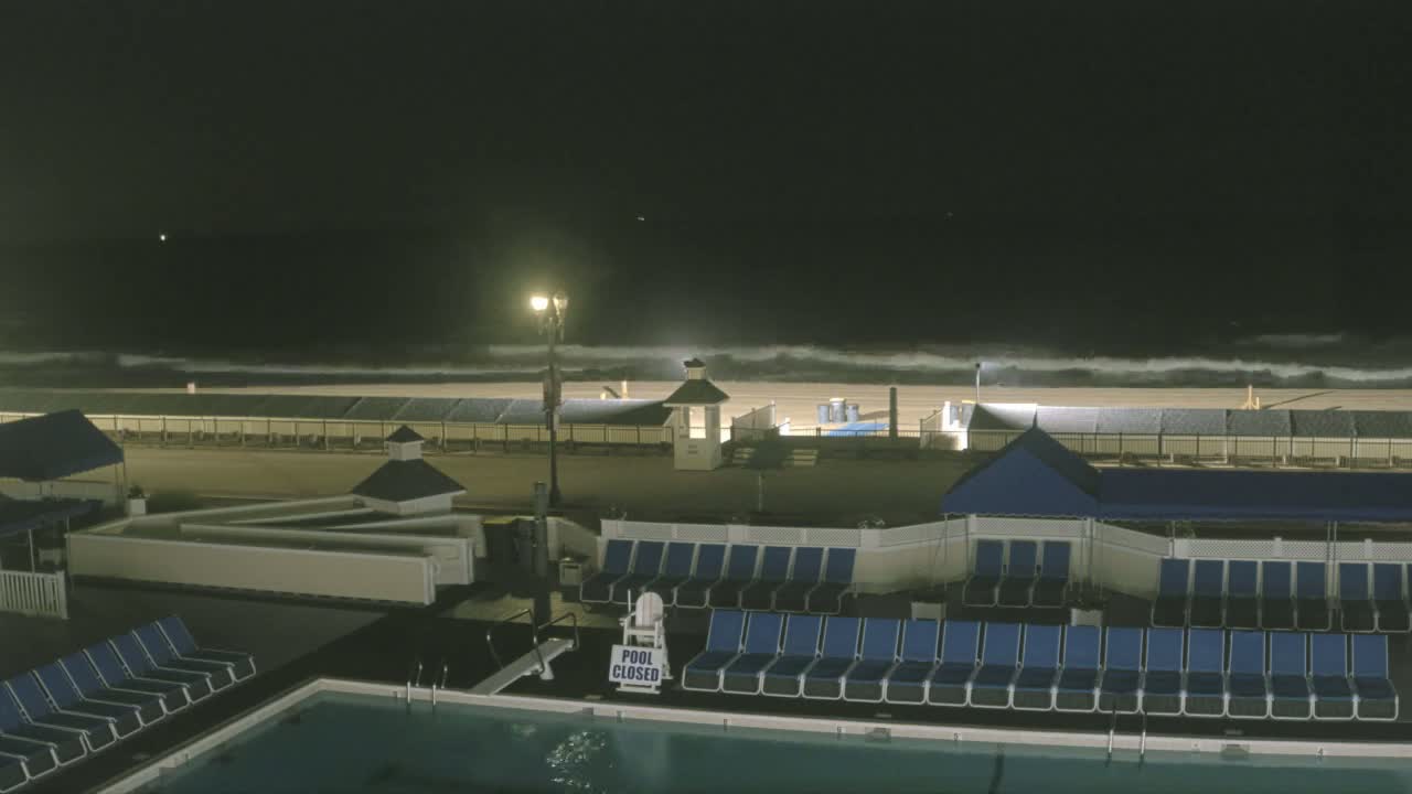 At night, a closed pool with rows of empty lounge chairs sits next to a beach with moderate waves under a dark sky.