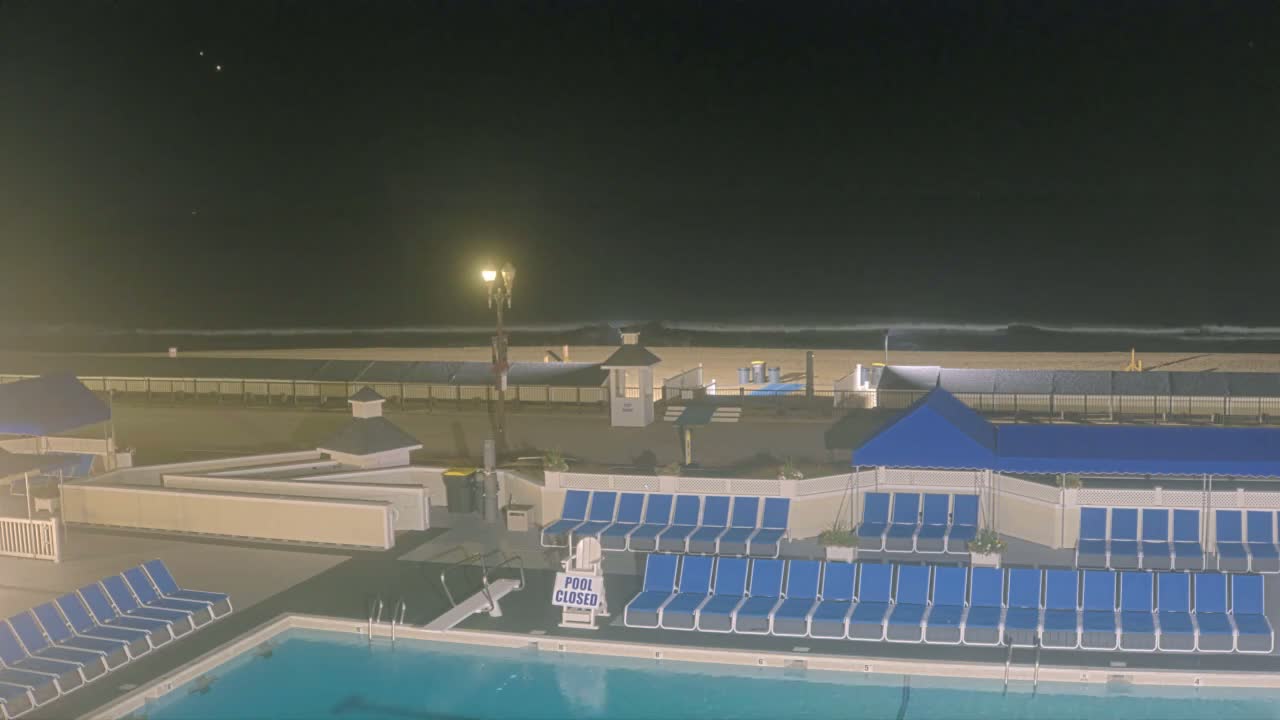 At night, a closed pool with rows of blue lounge chairs is situated next to a beach with gently lapping waves under a dark sky.