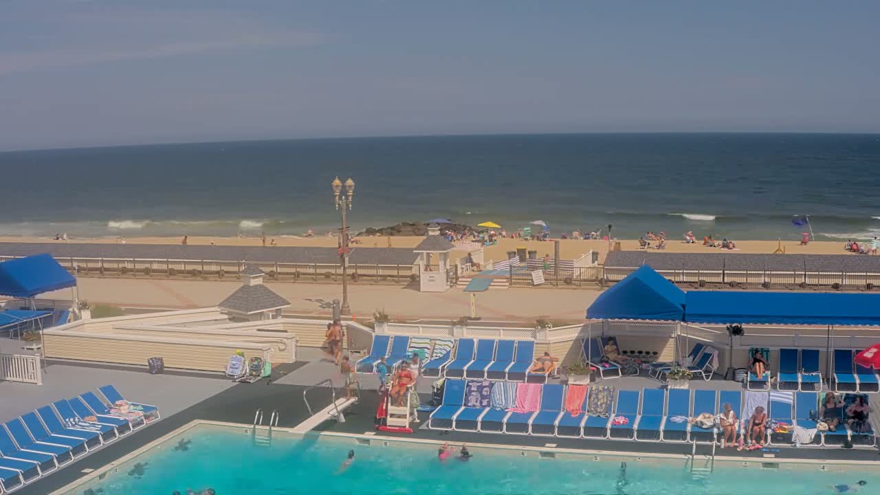 An outdoor swimming pool with people swimming and lounging on surrounding chairs overlooks a sunny beach with people relaxing and the ocean with small waves in the background.