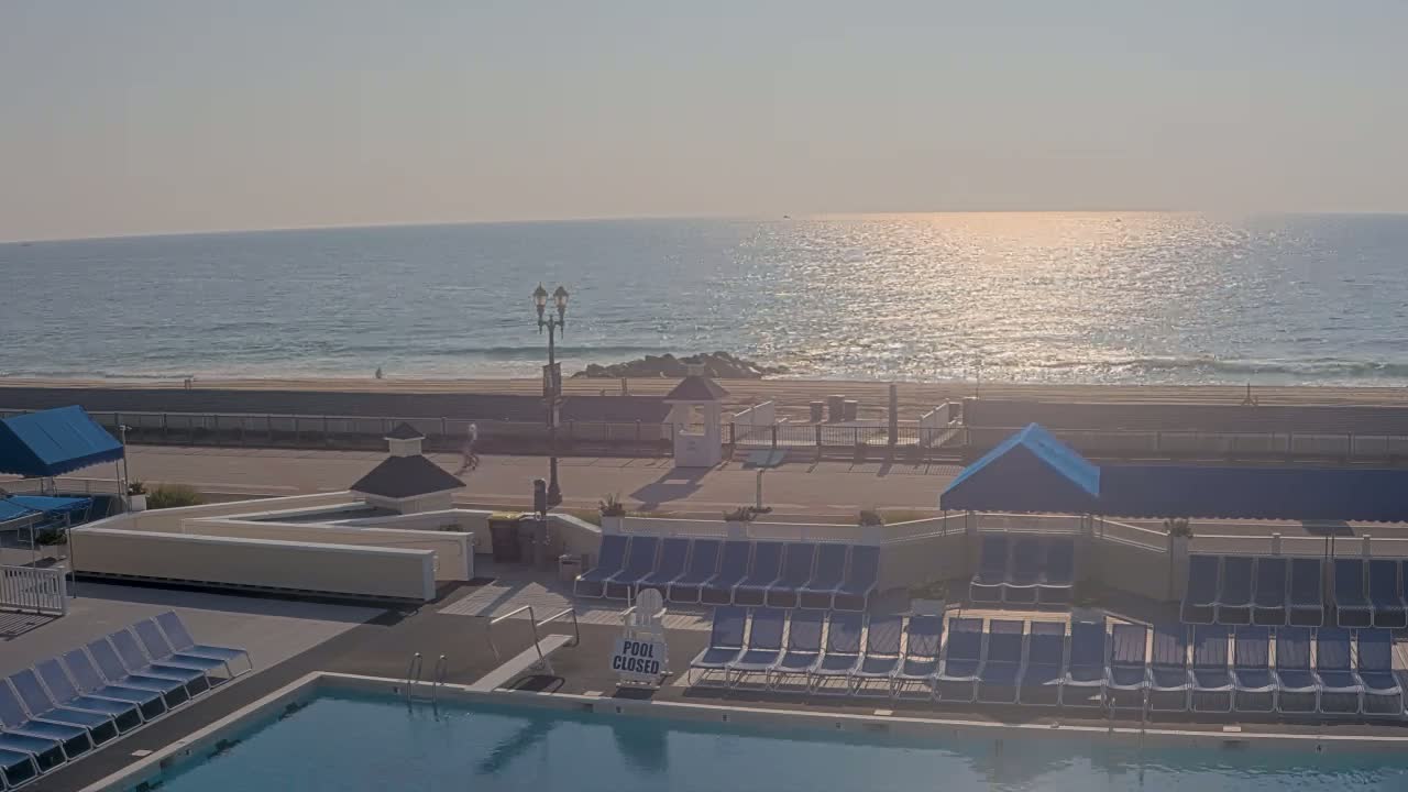 An outdoor swimming pool with people swimming and lounging on surrounding chairs overlooks a sunny beach with people relaxing and the ocean with small waves in the background.