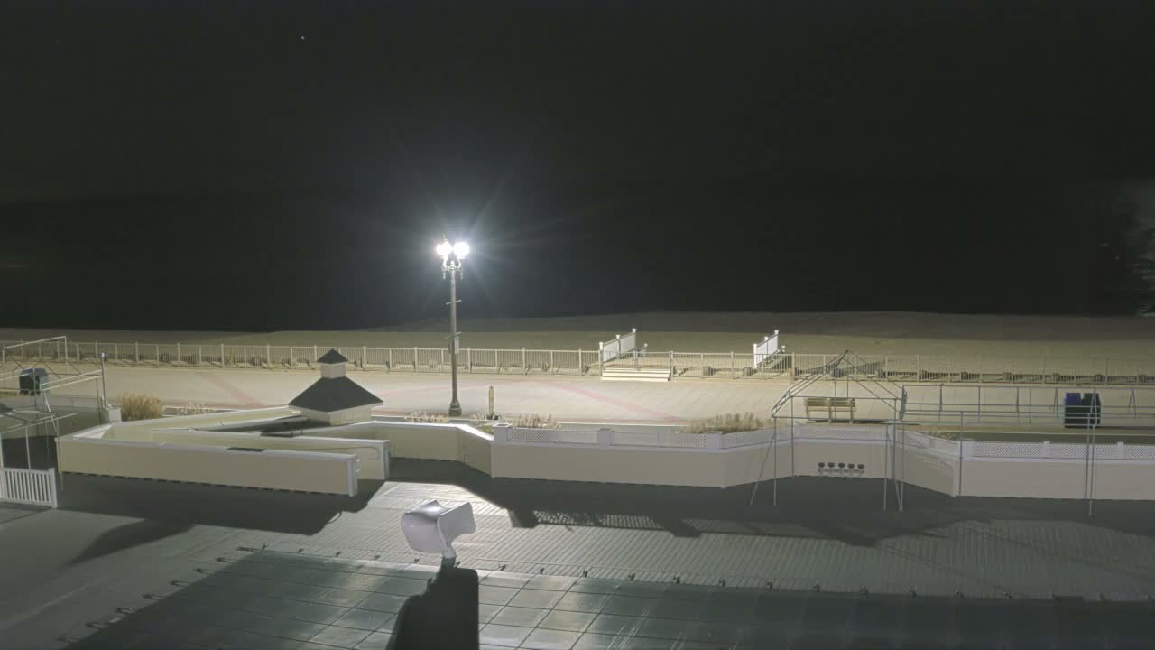 The image depicts an outdoor resort area at night, with a covered pool deck in the foreground, leading to a paved promenade featuring a small building and a bright lamppost, all bordered by a white fence along a sandy beach under a clear, dark sky.