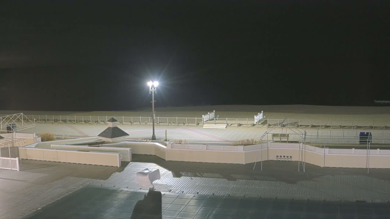 A brightly lit street lamp illuminates a deserted boardwalk with various low structures and fences, extending to a sandy beach and the dark ocean under a clear night sky.
