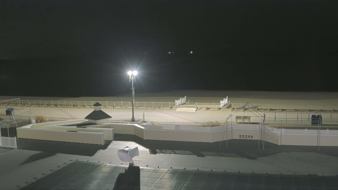 A clear night view captures a brightly lit boardwalk featuring a street lamp, gazebo, and benches, bordering a sandy beach enclosed by a white fence and steps.