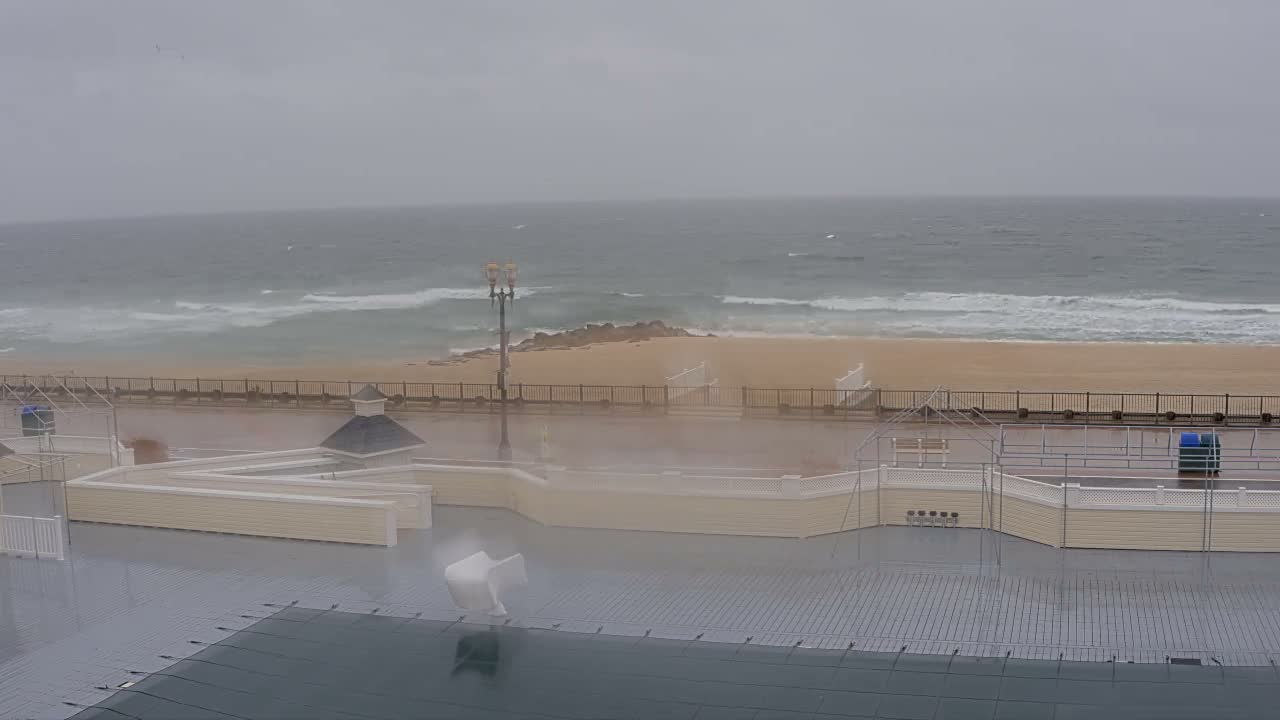 The scene shows a stormy, blustery day at the beach, with strong waves crashing on the sandy shore, a wet promenade, and light-colored structures in the foreground beneath a heavily overcast sky.