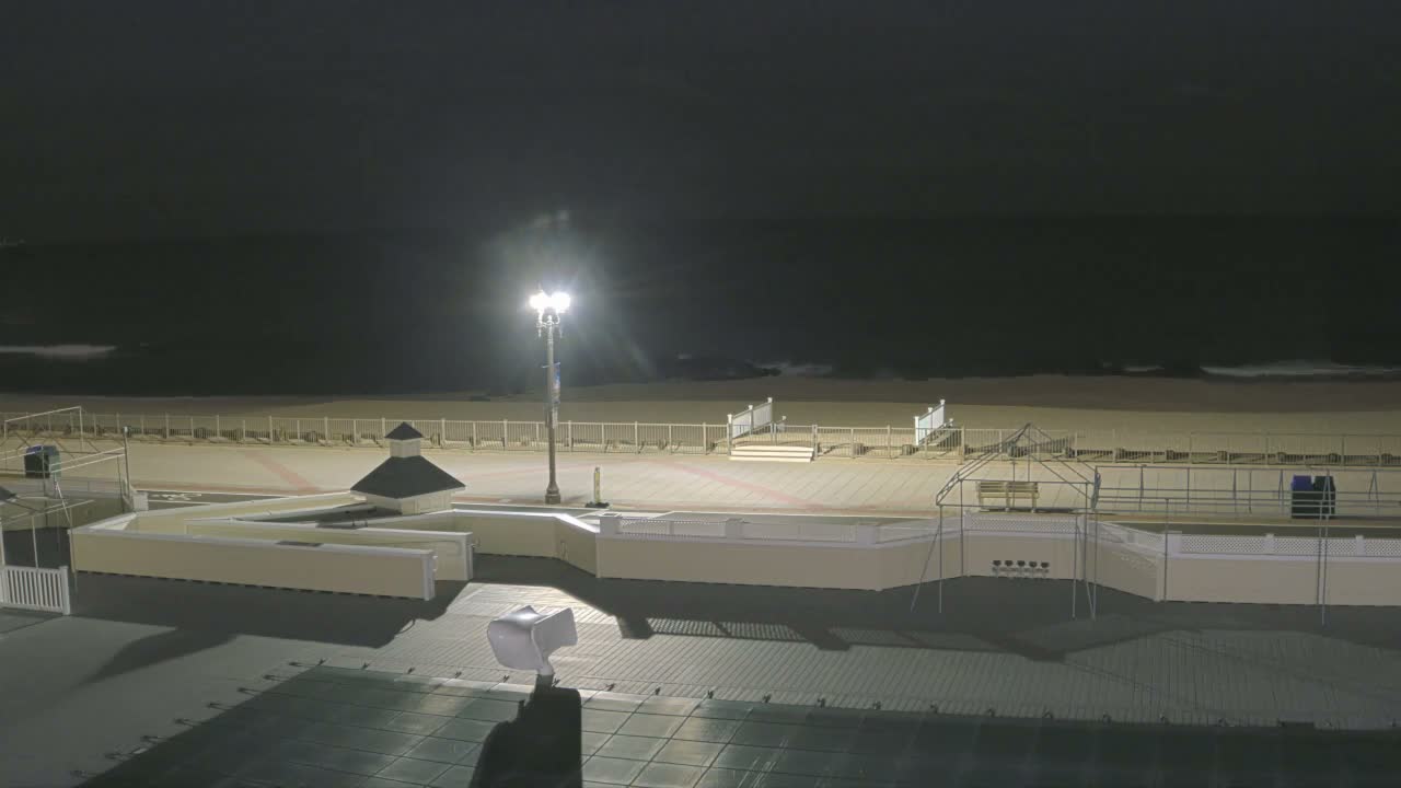 A tranquil nighttime scene displays a partially lit boardwalk and sandy beach alongside a dark ocean with visible waves, all under a clear night sky.