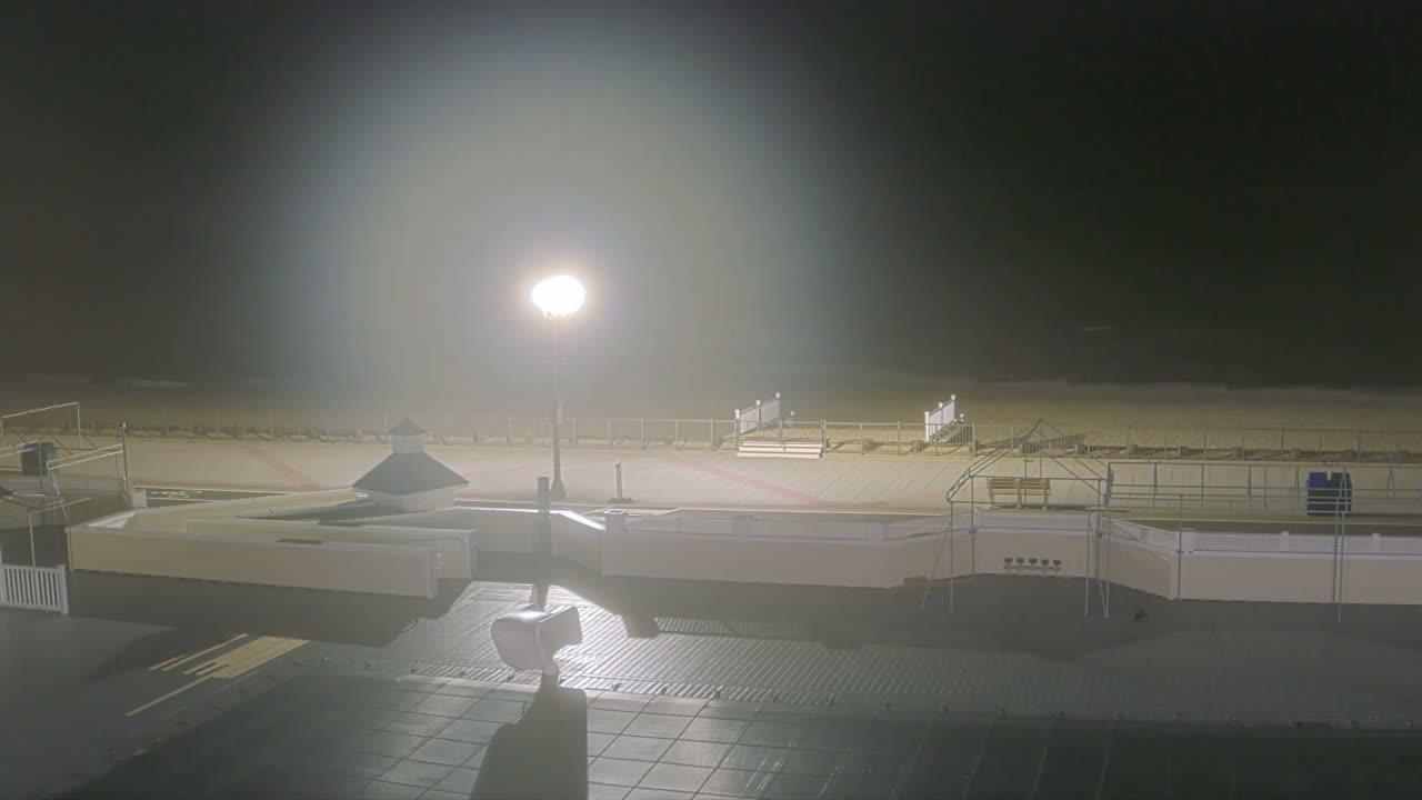 A tranquil nighttime scene displays a partially lit boardwalk and sandy beach alongside a dark ocean with visible waves, all under a clear night sky.