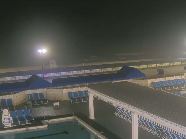 A nighttime view of a closed pool area with numerous blue lounge chairs under covered structures, overlooking a hazy beach under a dark sky.
