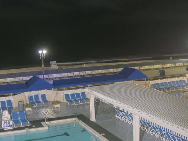 A nighttime view of a closed pool area with blue lounge chairs and umbrellas near an ocean under a dark sky.
