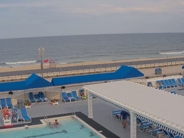 A nighttime view of a closed pool area with blue lounge chairs and umbrellas near an ocean under a dark sky.
