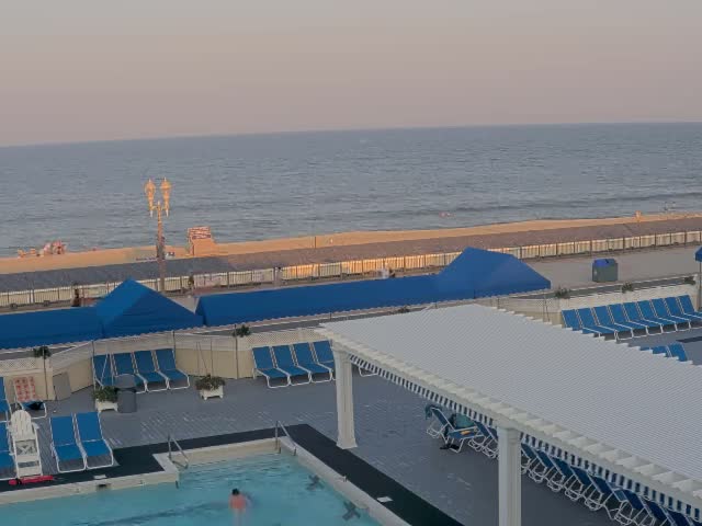 A nighttime view of a closed pool area with blue lounge chairs and umbrellas near an ocean under a dark sky.
