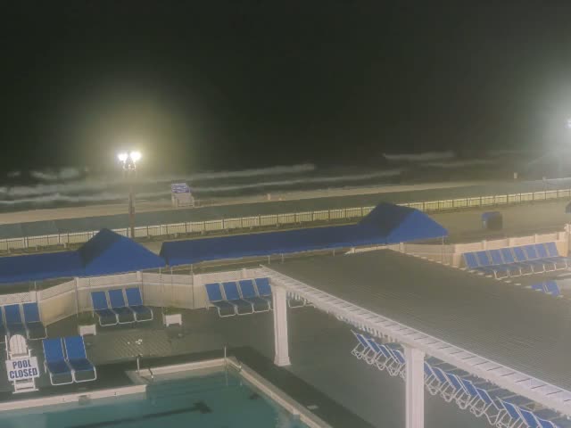 A nighttime view of a closed pool area with blue lounge chairs and cabanas next to a beach with moderate waves under a dark, cloudy sky.