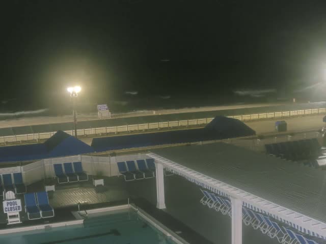 A nighttime view of a closed pool area with lounge chairs, next to a beach with choppy waves under a hazy sky.