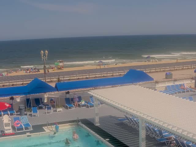 An outdoor pool area with lounge chairs and umbrellas overlooks a beach with people sunbathing and swimming in the ocean under a sunny sky.