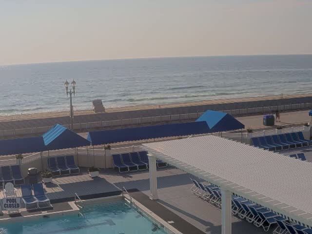 An outdoor pool area with lounge chairs and umbrellas overlooks a beach with people sunbathing and swimming in the ocean under a sunny sky.