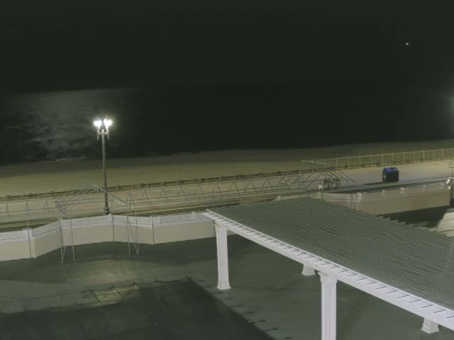 A clear night reveals a beach and ocean with gentle waves under a dark sky, illuminated by a prominent streetlight on a boardwalk and a long covered walkway extending into the foreground.