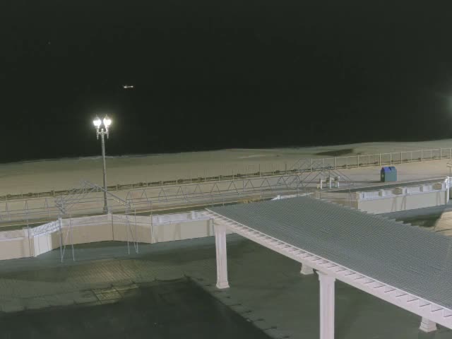 A clear night reveals a beach and ocean with gentle waves under a dark sky, illuminated by a prominent streetlight on a boardwalk and a long covered walkway extending into the foreground.
