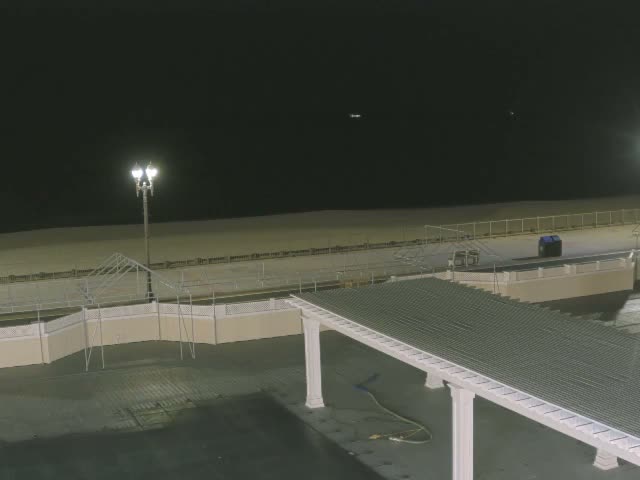 A clear night reveals a sandy beach with a fence and lamppost in the distance, while a pergola structure and railings are visible in the foreground.