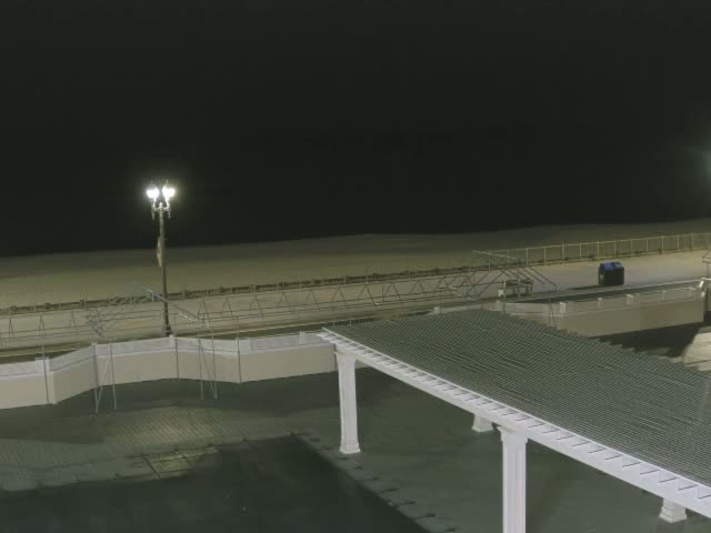 At night, a brightly lit streetlamp illuminates an empty beach and boardwalk alongside a covered walkway in the foreground, under clear skies.