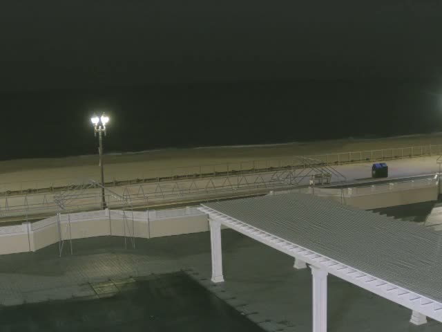 A clear night view reveals a dimly lit outdoor scene featuring a sandy beach meeting a dark ocean, a boardwalk with a bright street lamp and a blue bin, and a large white pergola extending into the foreground.