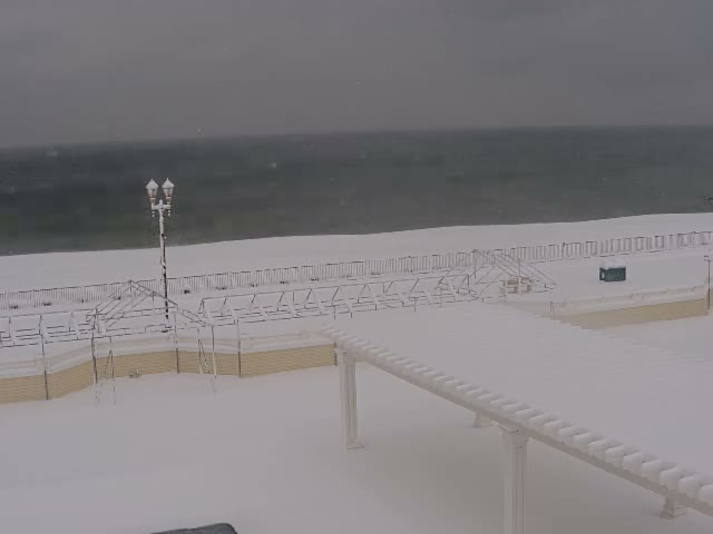 A clear night view reveals a dimly lit outdoor scene featuring a sandy beach meeting a dark ocean, a boardwalk with a bright street lamp and a blue bin, and a large white pergola extending into the foreground.