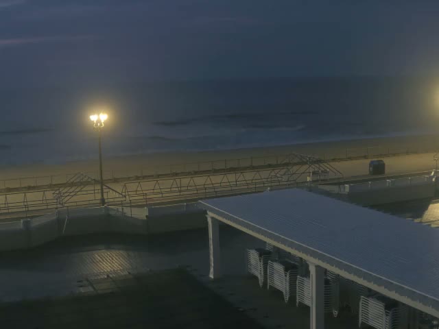 A clear night view reveals a dimly lit outdoor scene featuring a sandy beach meeting a dark ocean, a boardwalk with a bright street lamp and a blue bin, and a large white pergola extending into the foreground.