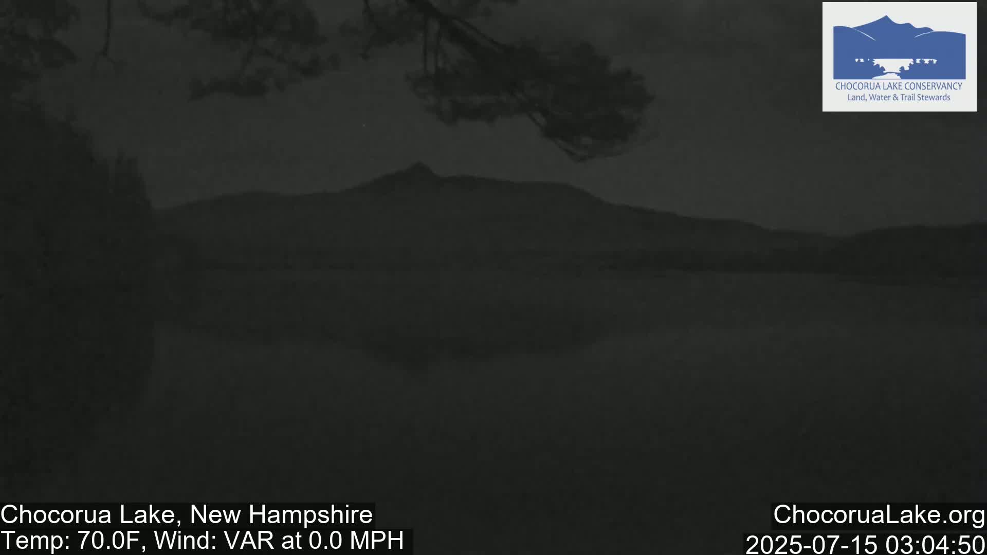 A dark, still lake is seen at dusk, with a mountain silhouetted in the distance under a clear sky.