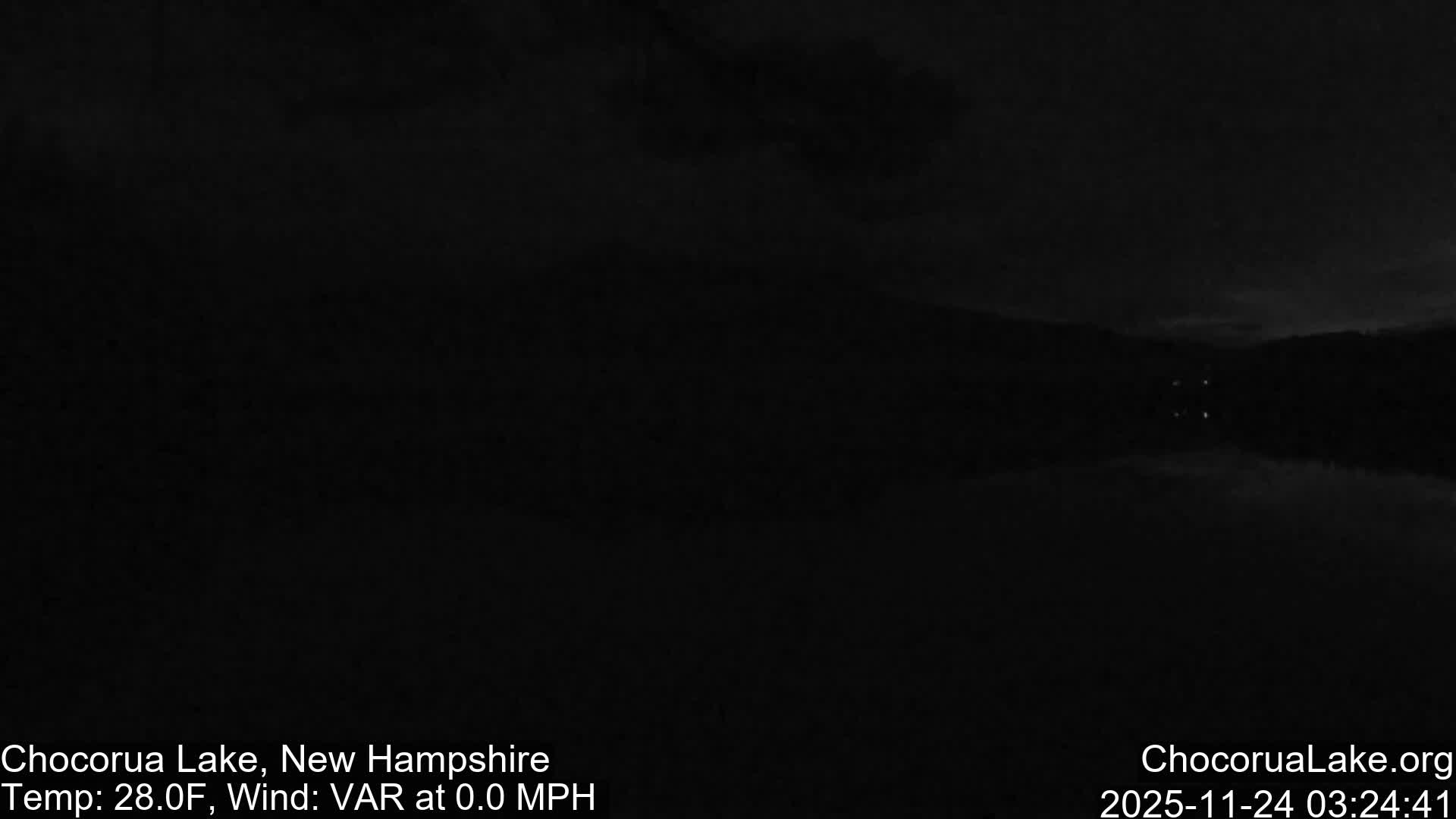 Under very dark nighttime conditions, a body of water reflects faint distant lights, with subtle outlines of land visible beneath a dark sky.