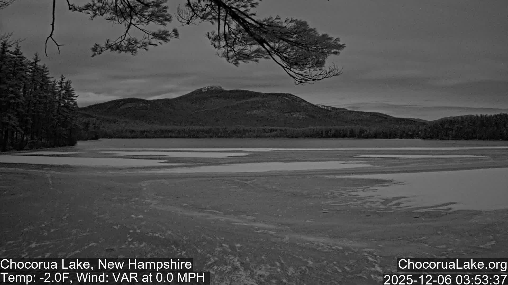 A monochromatic outdoor scene captures a partially frozen lake in the foreground, bordered by an evergreen forest and distant mountains under an overcast sky.