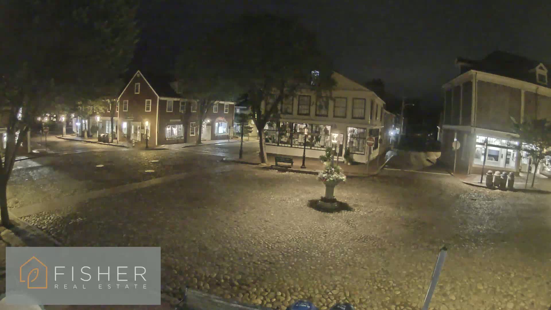 A cobblestone town square at night is illuminated by streetlights, showing several historic buildings surrounding it.