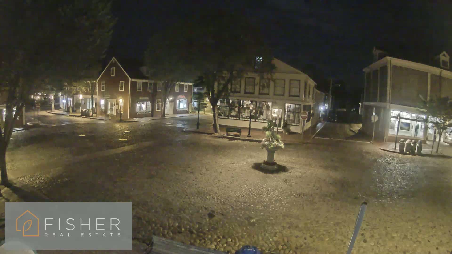 A cobblestone town square at night, illuminated by streetlights, is surrounded by several two-story buildings.