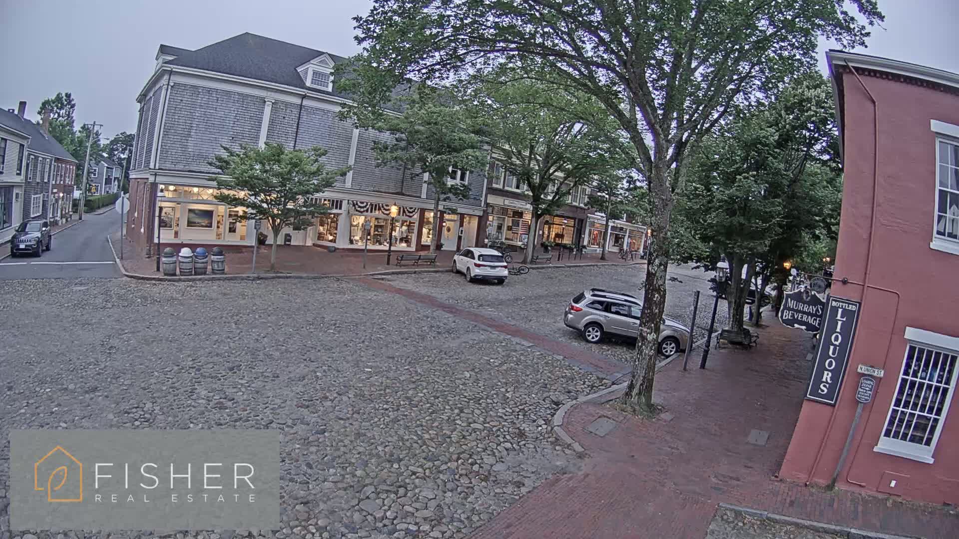 A nighttime street scene shows a cobblestone road lined with two-story buildings, a few parked cars, and trees under a dark sky.