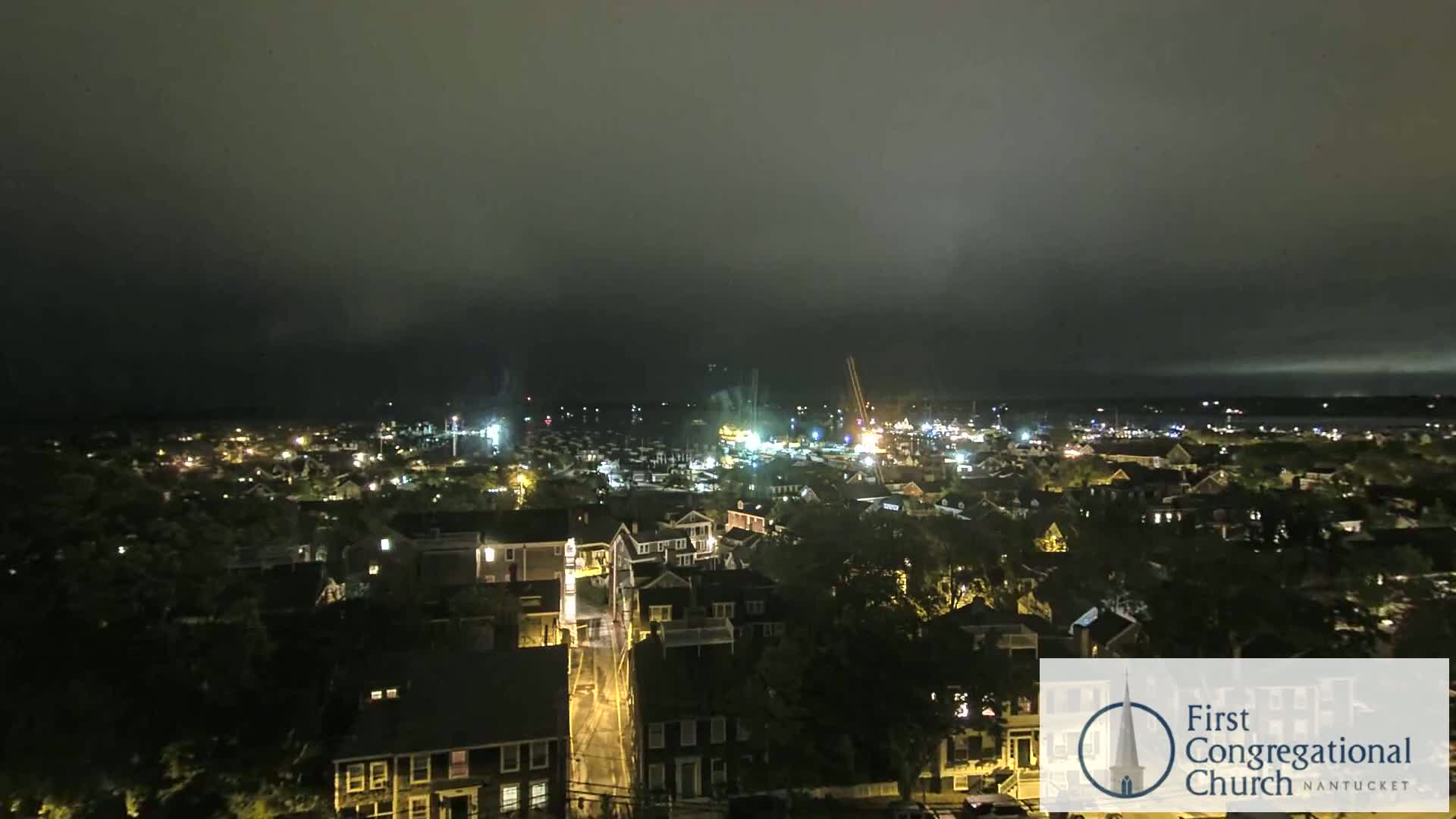 A nighttime view of a town illuminated by streetlights and house lights under an overcast sky.