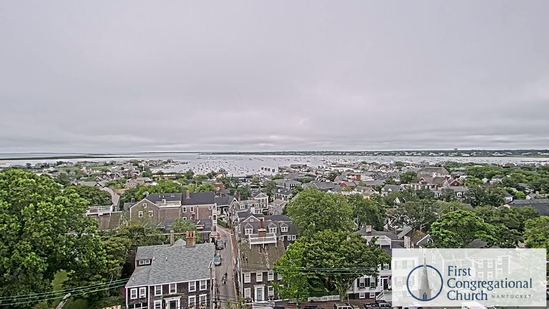 An elevated view of a coastal town on an overcast day shows a harbor filled with boats in the distance.