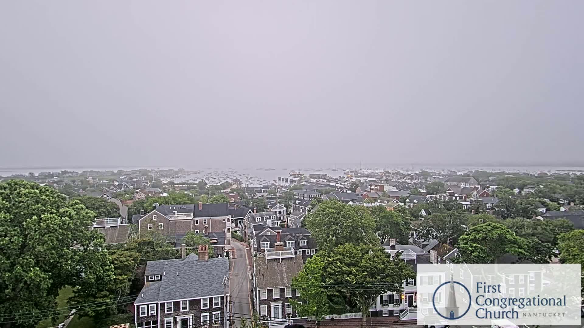 An elevated view of a coastal town on an overcast day shows a harbor filled with boats in the distance.