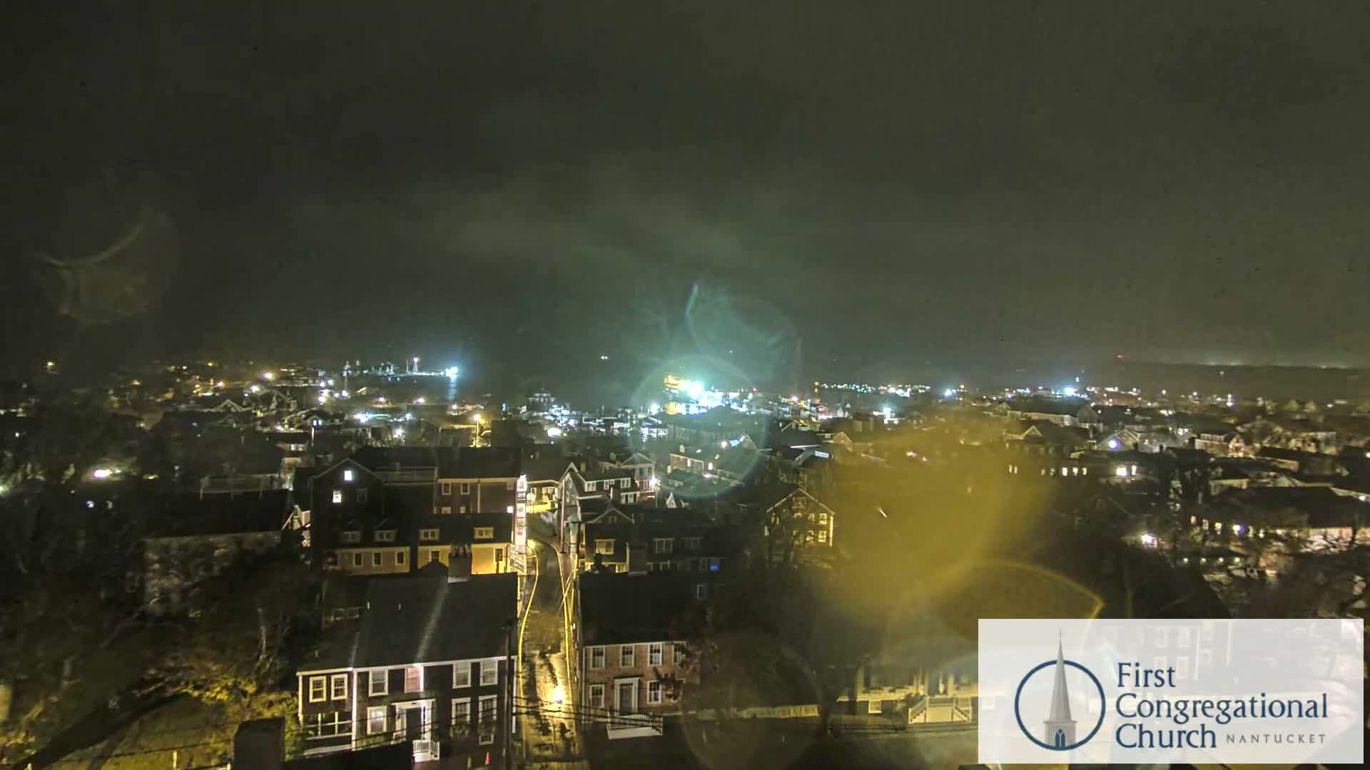 An overcast night captures a sprawling town illuminated by numerous streetlights and building windows, with a brightly lit waterfront visible in the distance.