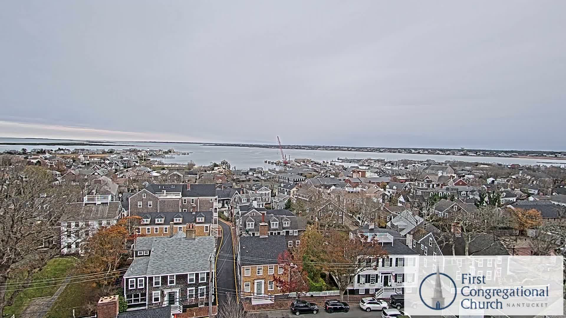 An overcast night captures a sprawling town illuminated by numerous streetlights and building windows, with a brightly lit waterfront visible in the distance.