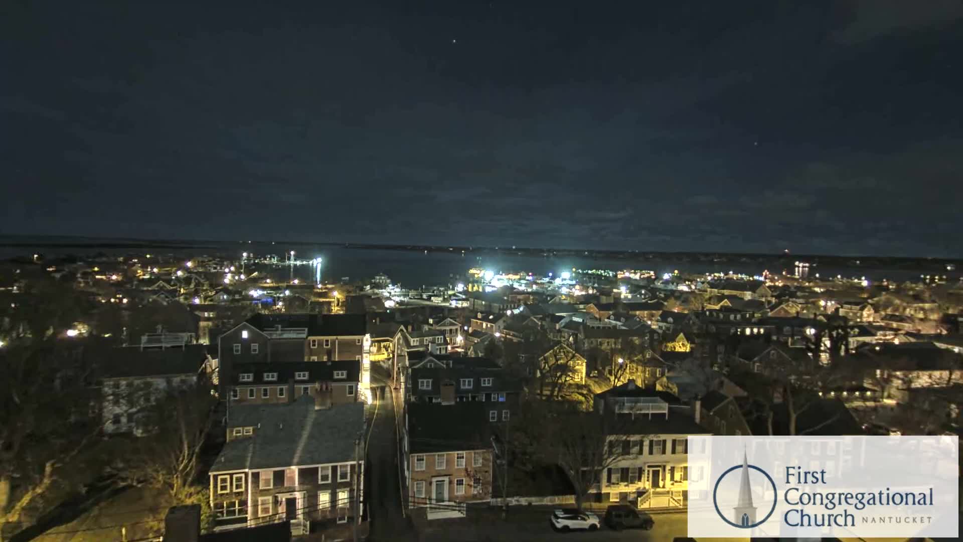 An aerial view reveals a coastal town illuminated at night under a partly cloudy sky, with numerous building and street lights glowing across the land and reflecting on the calm water.