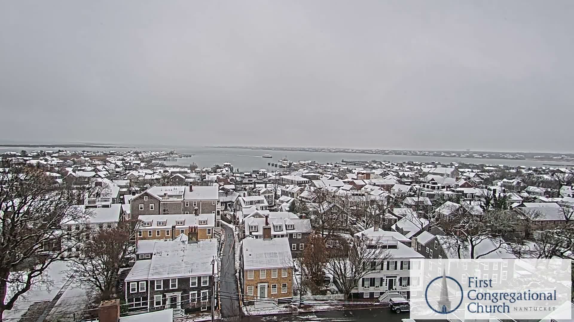 An aerial view reveals a coastal town illuminated at night under a partly cloudy sky, with numerous building and street lights glowing across the land and reflecting on the calm water.