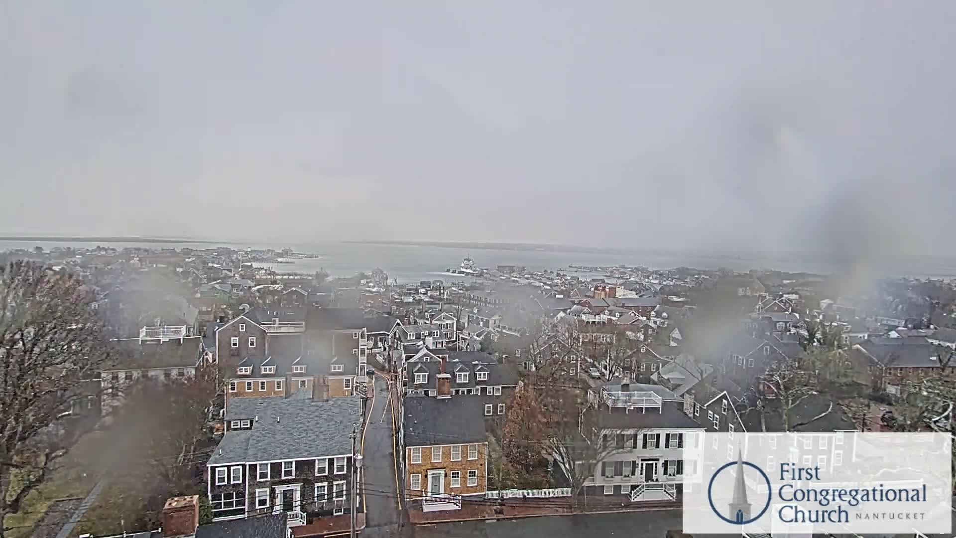 An aerial view reveals a coastal town illuminated at night under a partly cloudy sky, with numerous building and street lights glowing across the land and reflecting on the calm water.